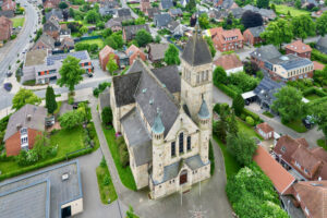 Luftaufnahme einer großen Steinkirche mit einem hohen zentralen Turm, umgeben von Wohnhäusern, grünen Bäumen und Straßen in einem Vorort. (Text mit KI erstellt)