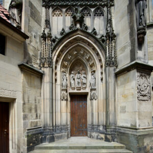 Eine gewölbte Holztür im gotischen Stil, eingebettet in eine kunstvolle Kirchenfassade aus Stein, verziert mit aufwendigen Schnitzereien, Statuen und Spitzbögen über dem Eingang. Eine Treppe führt hinauf zum Eingang. (Text mit KI erstellt)