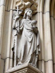 Eine steinerne Statue einer gewandeten Figur, die einen Stab in der einen und einen Kelch in der anderen Hand hält, steht in einer verzierten architektonischen Nische an einer Gebäudefassade. (Text mit KI erstellt)