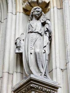 Eine steinerne Statue einer gewandeten Frau, die ein Buch und einen Stab in der Hand hält, steht in einer kunstvollen Nische an einer Gebäudefassade, und an ihrer Kleidung und der sie umgebenden Architektur sind komplizierte Details zu erkennen. (Text mit KI erstellt)