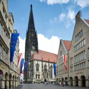 Eine gotische Kirche mit einem hohen, dunklen Turm steht auf einem europäischen Platz, flankiert von historischen Gebäuden, die mit der Europäischen Union und rot-gelben Fahnen geschmückt sind. Unter dem teilweise bewölkten Himmel spazieren Menschen über den offenen Platz. (Text mit KI erstellt)