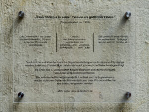 Eine durchsichtige Plakette mit deutschem Text, montiert auf einer strukturierten Steinwand. Der Text behandelt Jesus Christus in seiner Passion als göttlichen Erlöser und verweist auf die christliche und jüdische Symbolik, mit einem Website-Link am unteren Rand. (Text mit KI erstellt)