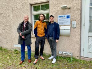 Drei Männer stehen vor einem beigen Gebäude in der Nähe eines Fensters und eines Schildes mit der Aufschrift Diakonie. Sie sind leger gekleidet und scheinen für ein Gruppenfoto auf einem kopfsteingepflasterten Weg zu posieren. (Text mit KI erstellt)