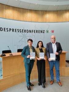 Drei Personen stehen vor der Kulisse einer Pressekonferenz und halten Broschüren in der Hand. Sie sind förmlich gekleidet und posieren für die Kamera. Im Hintergrund ist die PRESSEKONFERENZ NRW zu sehen, dahinter stehen ein Konferenztisch und Stühle. (Text mit KI erstellt)