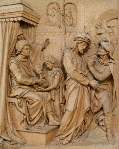Ein detailliertes Holzrelief stellt Pontius Pilatus dar, der sich die Hände wäscht, während Jesus, der eine Dornenkrone trägt, von einem Wächter präsentiert wird. Zwischen Pilatus und Jesus steht ein Kind vor einem Hintergrund aus geschnitzten Figuren. (Text mit KI erstellt)