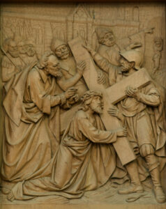 Ein detailliertes Holzrelief zeigt Jesus kniend und ein Kreuz tragend, umgeben von mehreren Figuren, von denen einige ihm beistehen und andere gestikulieren, vor einem architektonischen Hintergrund. (Text mit KI erstellt)
