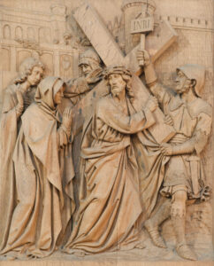 Ein detailliertes Holzrelief zeigt Jesus, der ein Kreuz trägt, umgeben von Trauernden und einem römischen Soldaten. Eine Person hält eine Plakette mit der Aufschrift INRI über Jesu Kopf, und im Hintergrund ist eine Stadtmauer zu sehen. (Text mit KI erstellt)
