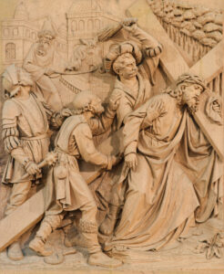 Eine detaillierte Holzschnitzerei zeigt Jesus, der ein Kreuz trägt, umgeben von Soldaten und Schaulustigen. Eine Figur erhebt eine Peitsche, während andere Jesus nach vorne schieben. Im Hintergrund sind architektonische Strukturen zu sehen. (Text mit KI erstellt)