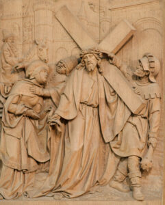 Ein detailliertes Holzrelief zeigt Jesus, der ein großes Kreuz trägt, umgeben von mehreren Figuren in historischer Kleidung, vor einem architektonischen Hintergrund. (Text mit KI erstellt)