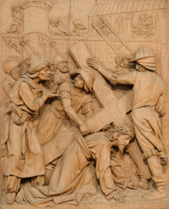 Ein geschnitztes Holzrelief zeigt Jesus, der ein großes Kreuz trägt und von mehreren Figuren umgeben ist; einige bieten ihre Hilfe an, während andere die Szene vor einem architektonischen Hintergrund betrachten oder lenken. (Text mit KI erstellt)