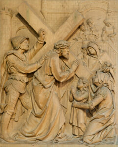 Ein detailliertes Holzrelief zeigt Jesus, der ein Kreuz trägt, umgeben von römischen Soldaten und Trauernden, darunter Frauen und ein Kind, das im Gebet kniet, und illustriert damit eine biblische Szene. (Text mit KI erstellt)