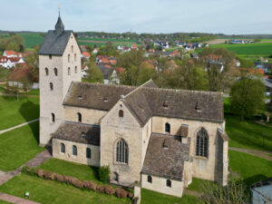 Luftaufnahme einer historischen Steinkirche mit einem hohen Glockenturm, umgeben von grünen Rasenflächen und Bäumen, mit einem Dorf und Feldern im Hintergrund. Die Kirche hat ein schräges Ziegeldach und Rundbogenfenster. (Text mit KI erstellt)