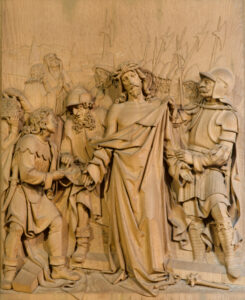 Das detaillierte Holzrelief zeigt den dornengekrönten Jesus, umgeben von römischen Soldaten und Männern mit Speeren, von denen einige sein Gewand halten und ihn verhöhnen, vor einer Kulisse von Schaulustigen. (Text mit KI erstellt)