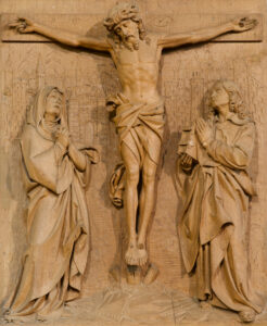 Ein detailliertes Holzrelief stellt die Kreuzigung Jesu dar, mit Jesus am Kreuz in der Mitte und zwei trauernden Figuren auf beiden Seiten, beide in betender Haltung. (Text mit KI erstellt)