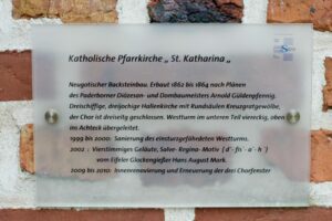 Eine Tafel an einer Backsteinmauer beschreibt die katholische Pfarrkirche St. Katharina in deutscher Sprache und gibt Auskunft über die neugotische Architektur, die Baugeschichte, die Renovierungen und die besonderen Merkmale. (Text mit KI erstellt)
