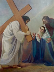 Ein Gemälde zeigt Jesus, der ein hölzernes Kreuz trägt und eine Dornenkrone trägt, während er einer trauernden Frau in einem blau-weißen Gewand, möglicherweise Maria, begegnet, die von einer anderen Figur gestützt wird. (Text mit KI erstellt)