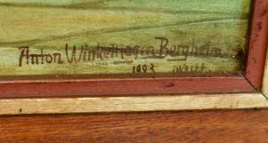 Nahaufnahme einer Gemäldesignatur in brauner Tinte mit der Aufschrift "Anton Winterberger Berghausen 1892" auf grünem Hintergrund, eingefasst von einem rot und beige gestrichenen Holzrahmen. (Text mit KI erstellt)