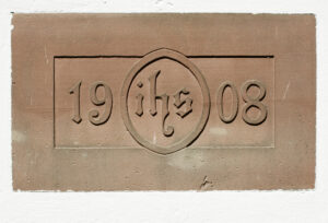 Eine rechteckige Steintafel mit der eingravierten Jahreszahl 1908, geteilt durch die Buchstaben ihs in einem Kreis, vor einer weißen Wand. (Text mit KI erstellt)