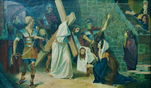 Ein Gemälde zeigt Jesus, der ein Kreuz trägt, umgeben von römischen Soldaten und Trauernden. Eine Frau kniet mit einem Tuch in der Hand vor ihm, während andere Figuren von nahe gelegenen Stufen und einer gewölbten Steinmauer aus zusehen. (Text mit KI erstellt)