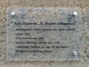 Eine Tafel an einer Steinmauer beschreibt die katholische Pfarrkirche St. Johannes Enthauptung, ihre Geschichte, Renovierungen und Besonderheiten wie einen Barockaltar von 1708 und ein romanisches Taufbecken. (Text mit KI erstellt)