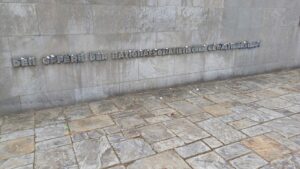 Gedenkmauer mit deutscher Aufschrift "DEN OPFERN DER NATIONALSOZIALISTISCHEN GEWALTHERRSCHAFT", davor Kieselsteine als Zeichen des Gedenkens auf den Boden gelegt. (Text mit KI erstellt)
