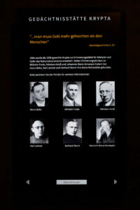Eine Tafel mit dem Titel Gedenkstätte Krypta zeigt Schwarz-Weiß-Porträts und die Namen von sechs Personen, mit einem deutschen Text zu Ehren der Opfer des Nationalsozialismus und einem Zitat aus Apostelgeschichte 5,29 am oberen Rand. (Text mit KI erstellt)
