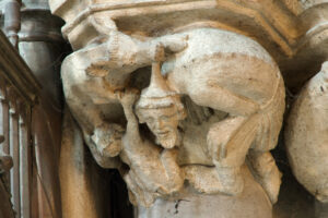 Ein gemeißeltes Steinkapitell, das eine menschliche Figur darstellt, die unter dem Gewicht eines gehörnten, tierähnlichen Wesens kauert, mit detaillierten Gesichtszügen und antiken Architekturelementen. In den Ritzen sind Spinnweben sichtbar. (Text mit KI erstellt)