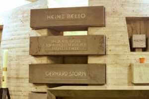 Betonwand mit eingravierten horizontalen Platten, auf denen Namen stehen: Heinz Bello, Nikolaus Gross Johannes Lauritzen Wilhelm Frede, Gerhard Storm. Links und rechts davon stehen eine Kerze und eine kleine Topfpflanze. (Text mit KI erstellt)