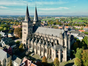 Luftaufnahme einer großen gotischen Kirche mit zwei Türmen, umgeben von kleinen Häusern und Bäumen in einer Stadt, mit offener Landschaft im Hintergrund unter blauem Himmel. (Text mit KI erstellt)
