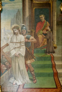 Ein Gemälde zeigt Jesus, der eine Dornenkrone und ein weißes Gewand trägt und von einem römischen Soldaten vor einen sitzenden Herrscher geführt wird, während eine Frau eine Schale präsentiert. Im Hintergrund sind eine Menschenmenge und ein großes Kreuz zu sehen. (Text mit KI erstellt)