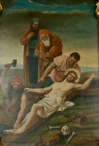 Ein Gemälde zeigt Jesus, der mit einer Dornenkrone auf dem Boden liegt und von zwei Männern an das Kreuz genagelt wird, während zwei andere zusehen. In der Nähe ruhen Knochen und ein Schädel unter einem bewölkten Himmel. (Text mit KI erstellt)