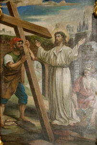 Ein Gemälde zeigt Jesus in einem weißen Gewand und einer Dornenkrone neben einem Mann, der ein großes Holzkreuz hält; im Hintergrund sind weitere Figuren und antike Stadtgebäude zu sehen. (Text mit KI erstellt)
