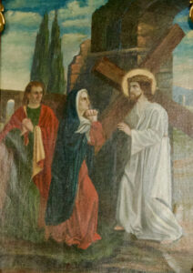 Ein Gemälde zeigt Jesus, der ein Holzkreuz trägt und ein weißes Gewand mit Heiligenschein trägt. Er begegnet einer trauernden Frau und einem Mann in bunten Gewändern, mit alten Steinruinen und Bäumen im Hintergrund. (Text mit KI erstellt)