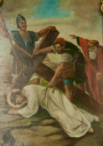 Ein Gemälde zeigt Jesus, der unter dem Gewicht eines großen Holzkreuzes zusammenbricht, mit Soldaten und einem älteren Mann in der Nähe. Ein Soldat hilft beim Anheben des Kreuzes, während der ältere Mann eine Geste nach vorne macht. Jesus trägt ein weißes Gewand und einen Heiligenschein. (Text mit KI erstellt)