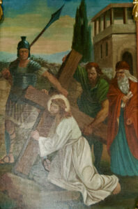 Ein Bild von Jesus, der ein Holzkreuz trägt und auf dem Boden kniet. Ein römischer Soldat steht mit einem Speer, während zwei Männer ihn stützen oder beobachten. Im Hintergrund sind ein Gebäude und Bäume zu sehen. (Text mit KI erstellt)