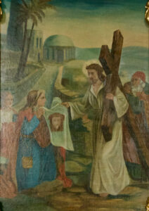 Ein Gemälde zeigt Jesus, der ein Holzkreuz trägt und einer Frau begegnet, die ein Tuch mit seinem Gesicht hält, während im Hintergrund weitere Figuren und antike Gebäude zu sehen sind. (Text mit KI erstellt)