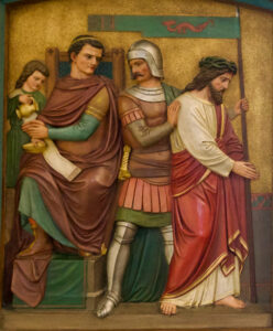 Ein detailliertes Relief zeigt Pontius Pilatus sitzend, der sich die Hände wäscht, einen Soldaten in Rüstung, der neben ihm steht, und Jesus, der eine Dornenkrone trägt und an den Händen gefesselt ist, der abgeführt wird; hinter Pilatus steht eine Dienerin mit einer Schale. (Text mit KI erstellt)