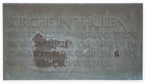 Eine Metalltafel mit der deutschen Aufschrift KIRCHE IN THÜLEN, die die Geschichte der ältesten Kirche Brilons beschreibt, mit teilweise abgenutztem und schwer lesbarem Text. (Text mit KI erstellt)