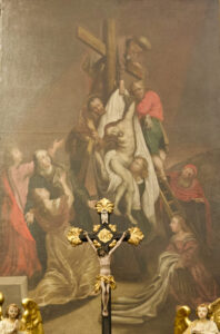 Ein klassisches Gemälde zeigt die Kreuzabnahme Jesu, der von mehreren Figuren vom Kreuz herabgelassen wird, während Trauernde zusehen; im Vordergrund steht ein Kruzifix mit zwei geschnitzten Engeln zu beiden Seiten. (Text mit KI erstellt)