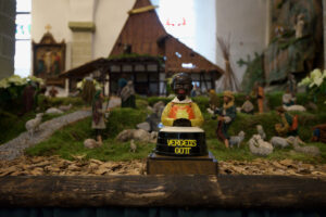 Eine dunkelhäutige Figur in einem gelben Gewand mit der Aufschrift VERGELTS GOTT steht vor einer Krippe mit verschiedenen Figuren, Tieren und einem rustikalen Stall im Hintergrund. (Text mit KI erstellt)