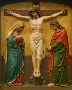 Eine Darstellung von Jesus am Kreuz mit zwei Figuren auf beiden Seiten, die beide Gewänder tragen und traurig dreinschauen. Der Hintergrund ist in einem gedämpften Goldton gehalten, und über dem Kopf Jesu steht INRI geschrieben. (Text mit KI erstellt)