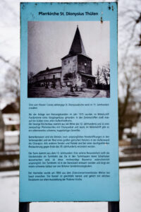 Eine historische Informationstafel über die St. Dionysius-Kirche in Thülen, mit einem Schwarz-Weiß-Foto der Kirche mit Steinturm und Spitzdach sowie einem beschreibenden deutschen Text darunter. (Text mit KI erstellt)