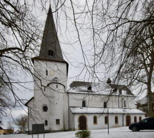 Eine weiße Kirche mit einem hohen, spitzen Kirchturm und einer Uhr steht an einem verschneiten Wintertag umgeben von blattlosen Bäumen. Ein schwarzes Auto ist in der Nähe geparkt, und der Boden ist leicht mit Schnee bedeckt. (Text mit KI erstellt)