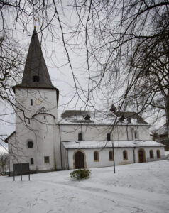 An einem verschneiten Wintertag steht eine weiße Kirche mit einem hohen Kirchturm inmitten kahler Bäume. Der Boden und das Dach sind mit Schnee bedeckt, und der Himmel ist bewölkt. (Text mit KI erstellt)
