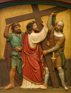 Eine Reliefskulptur, die Jesus darstellt, der ein großes Holzkreuz trägt, begleitet von zwei Männern - einer in Rüstung, der andere in einfacher Kleidung - vor einem bräunlichen Hintergrund. (Text mit KI erstellt)