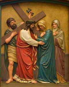 Eine Reliefskulptur zeigt Jesus, der ein Kreuz trägt und eine trauernde Frau in Blau und eine weitere Frau neben ihr tröstet, während ein Mann hinter Jesus steht und das Kreuz stützt. (Text mit KI erstellt)