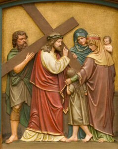 Eine Reliefskulptur zeigt Jesus, der ein Kreuz trägt und einer Gruppe von Frauen und Kindern begegnet. Eine Frau hält ein Kind, eine andere steht neben einem jungen Mädchen, das die Hand nach Jesus ausstreckt. Ein älterer Mann steht hinter Jesus und stützt das Kreuz. (Text mit KI erstellt)