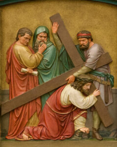 Ein Relief zeigt Jesus, der kniend ein großes Holzkreuz trägt, während drei Männer um ihn herumstehen; einer hält eine Schriftrolle, ein anderer gestikuliert nachdenklich, und der dritte stützt das Kreuz. (Text mit KI erstellt)