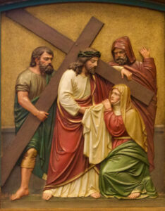 Eine Reliefskulptur zeigt Jesus, der ein Kreuz trägt und einer knienden Frau begegnet. Zwei weitere Männer stehen in der Nähe. Die Szene ist detailliert mit ausdrucksstarken Gesichtern und Gewändern in erdigen Farben gestaltet und stellt einen biblischen Moment dar. (Text mit KI erstellt)