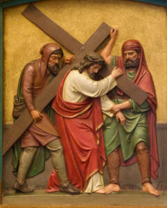 Eine Reliefskulptur, die Jesus zeigt, der ein großes Holzkreuz trägt, mit zwei Männern an seiner Seite. Jesus trägt eine Dornenkrone und ein rotes Gewand; ein Mann hilft, das Kreuz zu stützen, während der andere dicht hinter ihm geht. (Text mit KI erstellt)
